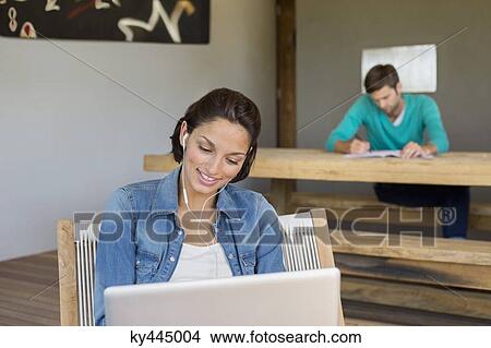 Woman listening to music on laptop with her husband in the background View Large Photo Image Picture - Woman listening to music on laptop with her husband in the background. Fotosearch