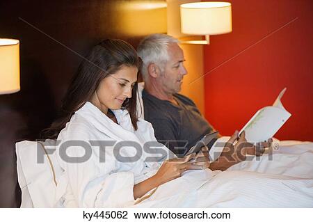 Woman using a digital tablet with her husband reading a book in a hotel room View Large Photo Image Stock Image - Woman using a digital tablet with her husband reading a book in a hotel room. Fotosearch