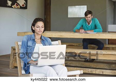Woman using a laptop with her husband in the background View Large Photo Image Stock Image - Woman using a laptop with her husband in the background. Fotosearch