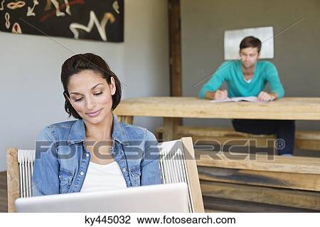 Stock Image - Woman using a laptop with her husband in the background. Fotosearch