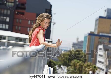 Stock Image - Smiling woman leaning against a railing and waving her hand. Fotosearch