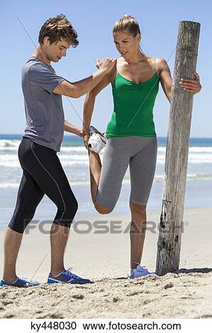 Stock Image - Man assisting a woman to exercise on the beach. Fotosearch