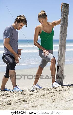 Stock Photograph - Man assisting a woman to exercise on the beach. Fotosearch