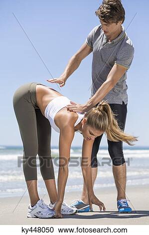 Stock Image - Man helping woman stretch at beach. Fotosearch