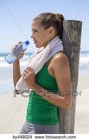 Woman leaning against a wooden post on the beach drinking water View Large Photo Image Picture - Woman leaning against a wooden post on the beach drinking water. Fotosearch