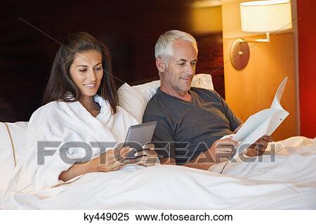 Woman using a digital tablet with her husband reading a book in a hotel room View Large Photo Image Stock Photography - Woman using a digital tablet with her husband reading a book in a hotel room. Fotosearch