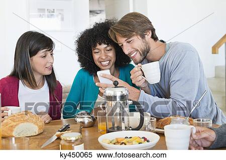 Friends sitting at a dining table and looking at a mobile phone View Large Photo Image Stock Photography - Friends sitting at a dining table and looking at a mobile phone. Fotosearch