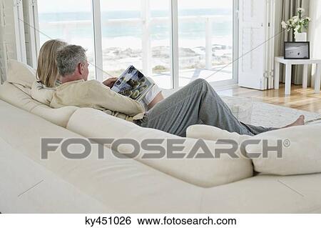 Couple sitting on a couch and reading a magazine View Large Photo Image Stock Photograph - Couple sitting on a couch and reading a magazine. Fotosearch