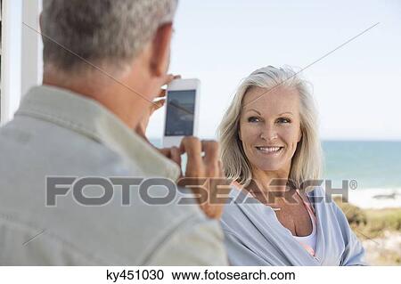 Man taking a picture of his wife with a cell phone on the beach View Large Photo Image Stock Image - Man taking a picture of his wife with a cell phone on the beach. Fotosearch