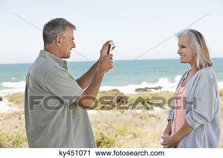 Man taking a picture of his wife with a cell phone on the beach View Large Photo Image Stock Image - Man taking a picture of his wife with a cell phone on the beach. Fotosearch