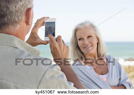 Stock Photo - Man taking a picture of his wife with a cell phone on the beach. Fotosearch