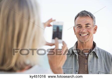 Woman taking a picture of her husband with a cell phone on the beach View Large Photo Image Stock Photograph - Woman taking a picture of her husband with a cell phone on the beach. Fotosearch