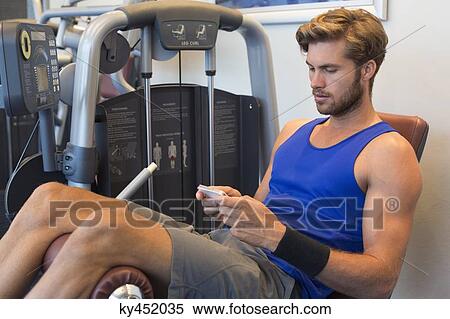Man sitting on an exercise machine and using a mobile phone in a gym View Large Photo Image Stock Photography - Man sitting on an exercise machine and using a mobile phone in a gym. Fotosearch