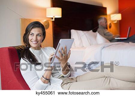 Woman using a digital tablet in a hotel room with her husband using laptop in the background View Large Photo Image Stock Image - Woman using a digital tablet in a hotel room with her husband using laptop in the background. Fotosearch