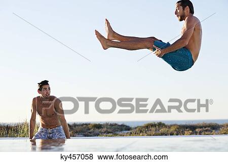 Stock Photo - Man jumping into a swimming pool with his friend standing at the poolside. Fotosearch