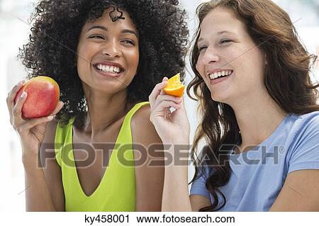 Stock Image - Close-up of two smiling female friends eating fruits. Fotosearch