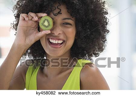 Stock Image - Smiling woman holding a kiwi fruit in front of her eye. Fotosearch