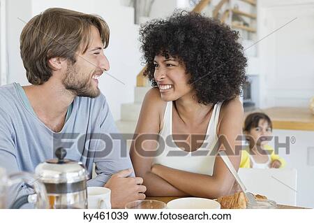 Couple smiling at a dining table with their daughter in the background View Large Photo Image Stock Photo - Couple smiling at a dining table with their daughter in the background. Fotosearch