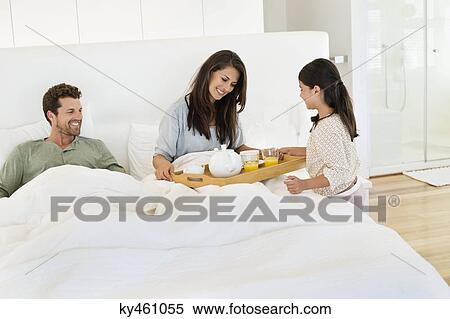Stock Photography - Girl serving tea to her parents on the bed. Fotosearch