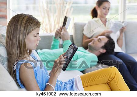 Stock Image - Girl using a digital tablet with her parents reading books at home. Fotosearch