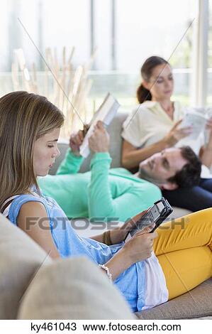 Stock Image - Girl using a digital tablet with her parents reading books at home. Fotosearch