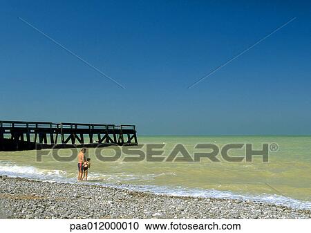 Stock Image - Father and daughter standing on beach at edge of water, near pier. Fotosearch