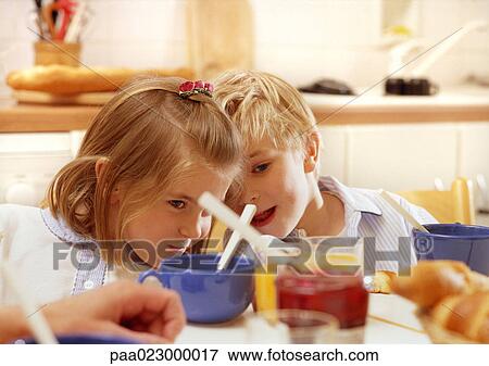 Stock Photo - Children sitting at table leaning heads together, head and shoulders, cups, jelly jar and adult hand blurred in foreground. Fotosearch