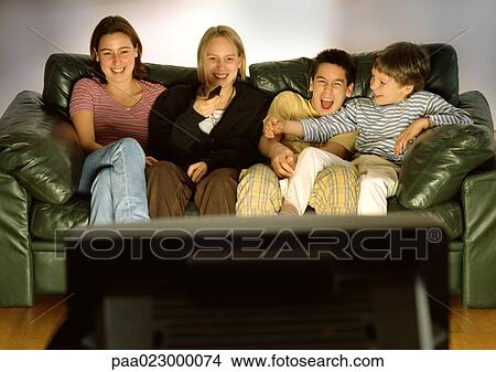 Picture - Three young people and one child sitting on green couch together, laughing, blurred rear view of tv in foreground. Fotosearch