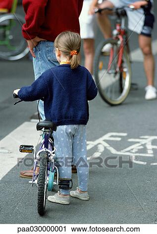 Stock Image - Young girl standing with bike near man and other bikers, view from behind.. Fotosearch