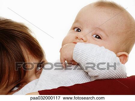 Stock Photo - Baby with hand in mouth looking at camera, woman, upper part of head, looking up at baby, close-up. Fotosearch