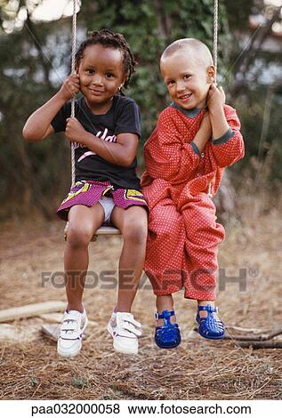 Stock Photo - Little boy and girl sitting on swing together, each holding one side of swing rope, smiling at camera, full length.. Fotosearch