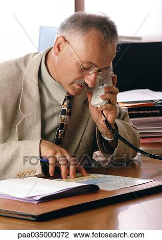 Stock Image - Man looking at documents while using phone. Fotosearch