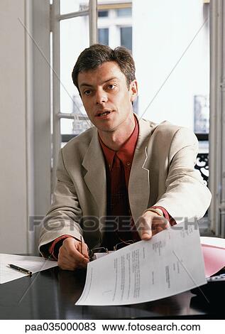 Stock Image - Man sitting at desk, holding document. Fotosearch