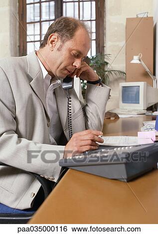 Stock Photograph - Man sitting at desk, telephoning, side view. Fotosearch