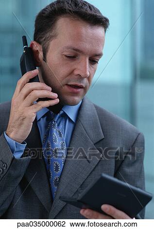 Stock Image - Man using cell phone while looking at pocket computer, portrait. Fotosearch