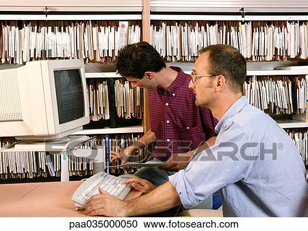 Stock Image - Man using computer, other man looking at files on shelves. Fotosearch