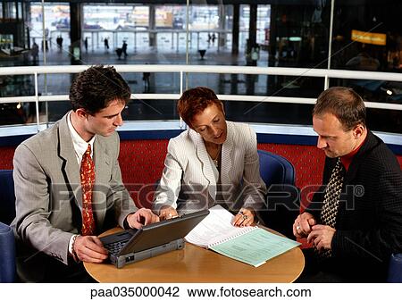 Stock Image - Three people sitting at table, one holding laptop computer. Fotosearch