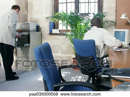 Stock Photo - Two men in a room, one using computer, rear view. Fotosearch