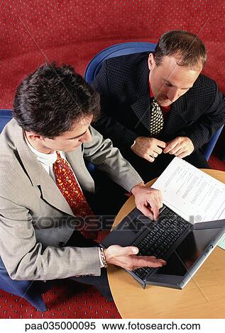 Stock Photography - Two men sitting at table, looking at laptop computer, elevated view. Fotosearch