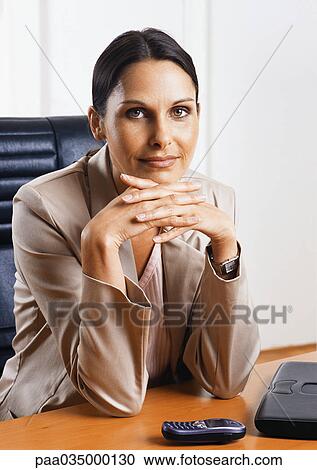Stock Image - Woman sitting at desk, hands together, portrait. Fotosearch