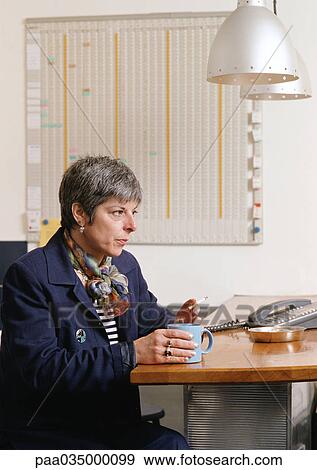 Stock Photo - Woman sitting at table, holding cup and smoking cigarette. Fotosearch