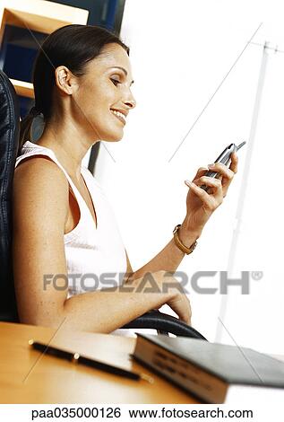 Stock Photograph - Woman sitting in armchair, looking at cell phone, side view. Fotosearch