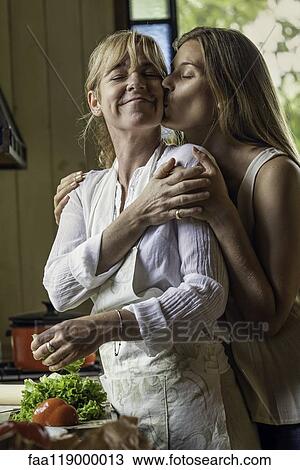 Stock Image - Daughter kissing her mother in kitchen. Fotosearch
