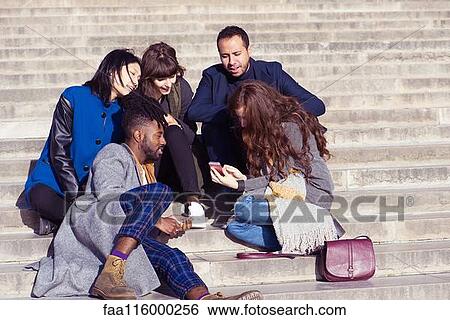 Stock Photograph - Friends sitting on steps and using smartphone. Fotosearch