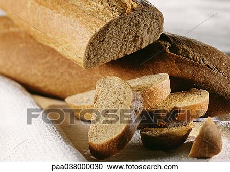 Stock Image - Two loaves of whole wheat bread, one whole, one cut, with slices of bread in a pile, close-up. Fotosearch