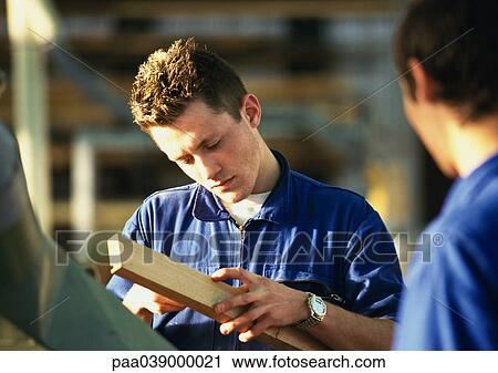 Stock Image - Man in blue coveralls holding plank of wood, second man rear view, blurred in foreground. Fotosearch