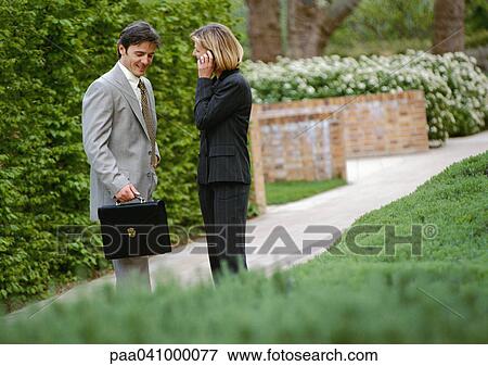 Stock Photo - Businessman and businesswoman standing in park, businesswoman using cell phone. Fotosearch