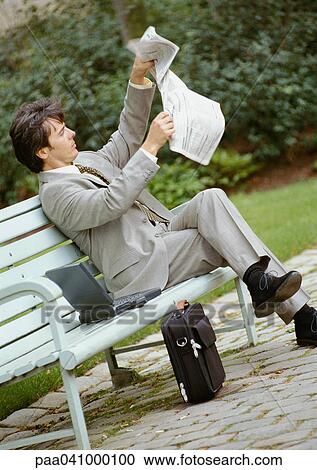 Stock Image - Businessman sitting on park bench, opening newspaper. Fotosearch