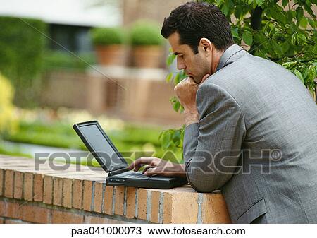 Businessman using laptop computer outside, leaning against low brick wall, side view View Large Photo Image Stock Image - Businessman using laptop computer outside, leaning against low brick wall, side view. Fotosearch
