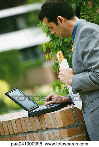 Businessman using laptop computer outside, holding sandwich View Large Photo Image Stock Photo - Businessman using laptop computer outside, holding sandwich. Fotosearch